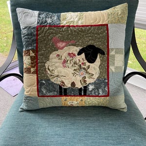 May include: A square quilted throw pillow featuring a central applique of a sheep with a floral patterned body and a solid black head. A small pink bird sits on the sheep's back. The center panel is framed by a red border and various patchwork squares.