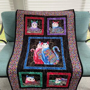 May include: A rectangular quilt featuring a black background with five framed panels depicting stylized cats in vibrant colors. The central panel shows two cats, one pink and one blue, surrounded by smaller panels with individual cat portraits.