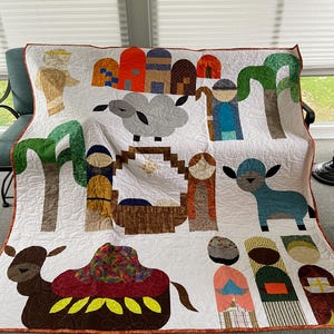 Pode incluir: Uma colcha branca apresentando uma cena de natividade com figuras aplicadas, incluindo um camelo, ovelhas, palmeiras e pessoas em tecidos coloridos com padrões. O design inclui uma manjedoura central, uma estrela e formas arquitetónicas em laranja, azul e castanho.