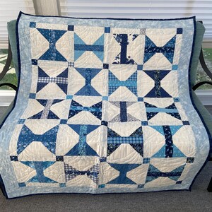 May include: A handmade quilt featuring a repeating bow-tie block pattern in various shades of blue and white. The design incorporates diverse blue fabric prints, including plaids, florals, and solids, set against a white background with a light blue border.