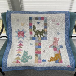 May include: A square quilt featuring a white background with a blue floral border. The design includes patchwork columns, a star, triangles, and several cat silhouettes in various floral fabrics. Small heart and cloud shapes are appliqued throughout.