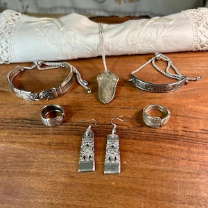 May include: A collection of vintage silver jewelry, including two bracelets, a pendant necklace, two rings, and a pair of earrings. The jewelry is displayed on a wooden surface with a white cloth in the background. The pieces have intricate designs.