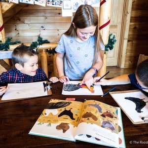 May include: Three children are sitting at a wooden table, drawing and coloring in a book about chickens. The book is open to a page with illustrations of different types of chickens. The children are using pencils and crayons to color the pictures.