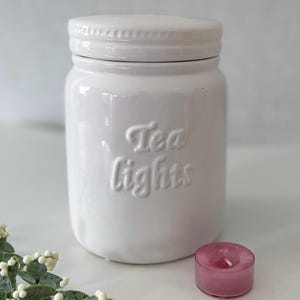 May include: White ceramic jar with a lid, labelled "Tea Lights". A pink tea light candle is in the foreground.