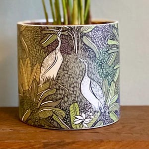 May include: A ceramic plant pot with a detailed illustration of white herons amidst lush green foliage. The design features various shades of green, brown, and white, creating a naturalistic scene. The pot sits on a wooden surface.