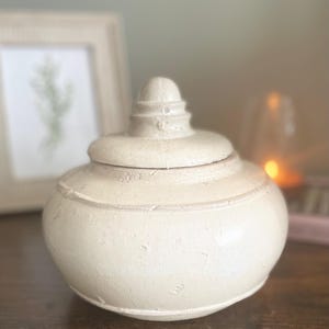 May include: A round, off-white ceramic jar with a textured, matte finish and a matching lid. The lid features a tiered, pointed finial at the top. The jar has a wide, bulbous body with a slightly tapered base and a distinct horizontal ridge.
