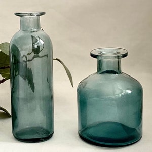 May include: Two glass vases in a teal blue colour. The vase on the left is taller and more cylindrical. The vase on the right is shorter and wider.