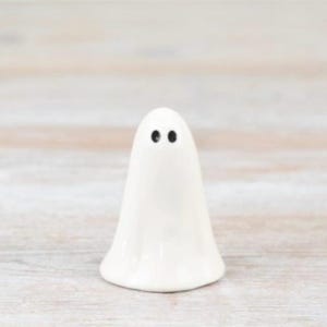 May include: A small, white ceramic ghost figurine with two black dot eyes. The ghost has a rounded base and a smooth, glossy finish. The figurine is standing on a light-coloured wooden surface.