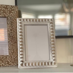 May include: Two decorative picture frames. One frame is white with a beaded border. The other frame is brown with a carved leaf design. Both frames have a white interior.