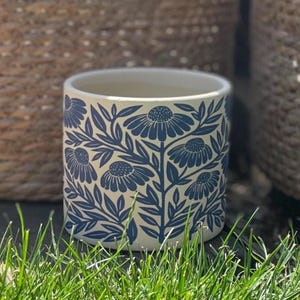 May include: A cylindrical ceramic plant pot with a cream base and a repeating pattern of blue floral designs. The pot is on green grass.