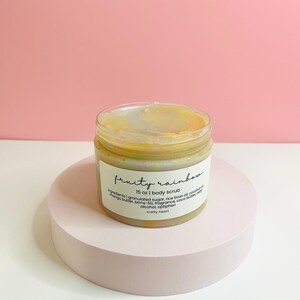 May include: A jar of "fruity rainbow" body scrub, 15 oz, sits on a light pink pedestal. The jar has a clear glass body with a white label. The label lists ingredients like granulated sugar, mango butter, and coconut oil.