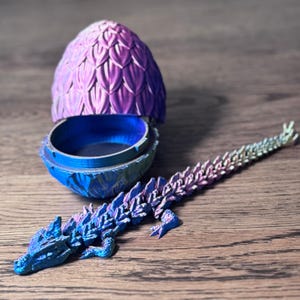 May include: A 3D-printed dragon egg with a matching articulated dragon. The egg is open, revealing a blue interior. The egg and dragon have a color-shifting finish, transitioning from blue to purple. The dragon is lying on a wooden surface.