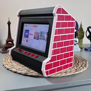 Op de afbeelding: Een zwart-witte arcade-console met een rood baksteenpatroon aan de zijkant. De console heeft een scherm met een game-interface, rode knoppen en een joystick. Het ontwerp doet denken aan klassieke arcadespellen.