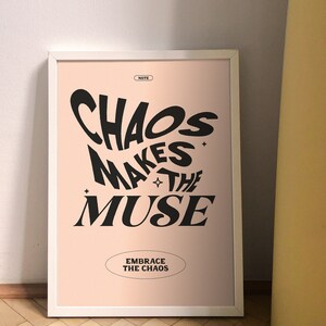 以下が含まれることがあります： ピンクの背景と黒い文字で「Chaos Makes the Muse」と書かれた額装されたプリント。その下に「Embrace the Chaos」と書かれた小さな楕円形があります。
