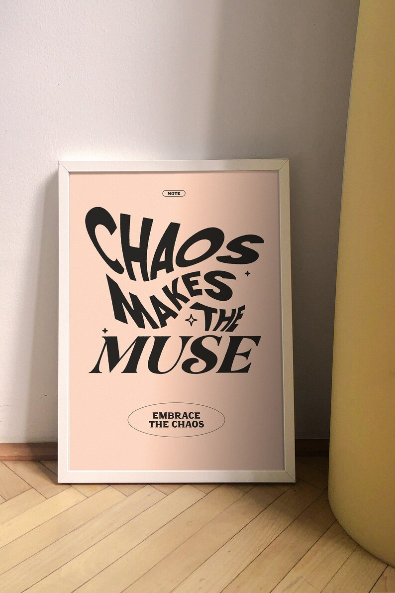 以下が含まれることがあります： ピンクの背景と黒い文字で「Chaos Makes the Muse」と書かれた額装されたプリント。その下に「Embrace the Chaos」と書かれた小さな楕円形があります。