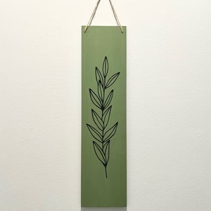 Puede incluir: Un letrero de madera verde con un dibujo de línea negra de una rama frondosa. El letrero está colgado de una cuerda.