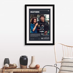 May include: A black framed movie poster for the film "Heathers" featuring a scene with two characters. The poster includes the film's title, release date, rating, and genre. The poster is on a white wall with a wooden table and a chair in the background.