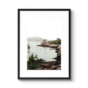 Puede incluir: Una fotografía en blanco y negro de una costa rocosa con un pequeño edificio de madera en la orilla. El agua está tranquila y el cielo está nublado.