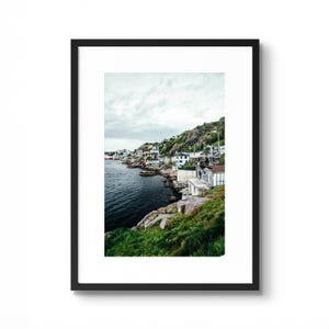 May include: A framed print featuring a coastal town nestled along a rocky shoreline. The image showcases a cluster of white and colorful buildings on a hillside, with dark blue water and green grass in the foreground. The sky is overcast.