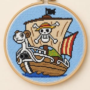 May include: A circular cross-stitch embroidery in a wooden hoop featuring a brown ship with a white ram figurehead. The sail displays a skull and crossbones wearing a yellow straw hat. The background is light blue with a small red and yellow flag.