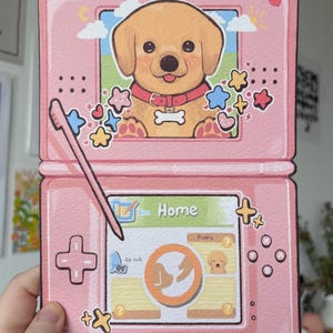 May include: A pink handheld gaming console illustration featuring a golden puppy on the top screen and a home menu interface on the bottom screen. A stylus is depicted resting diagonally across the device, surrounded by colorful star and heart icons.