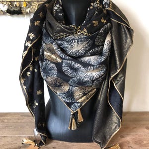 Peut inclure: Écharpe noire et dorée avec un motif floral et une bordure dorée, drapée sur un mannequin. Le tissu scintillant est orné de glands et de motifs de feuilles dorées.