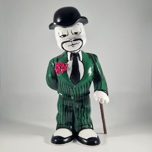 Puede incluir: Figura de cerámica de un hombre con traje verde a rayas finas, corbata negra y bombín. La figura tiene una cara blanca con bigote negro y sostiene un bastón marrón. Una flor roja está prendida en la solapa. Los zapatos son blancos y negros.