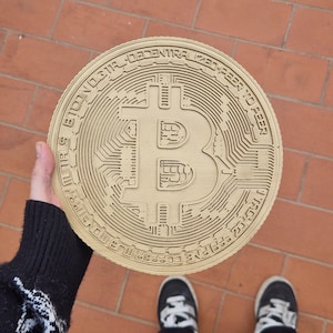 Puede incluir: Una moneda decorativa grande, redonda y de color beige con el logotipo de Bitcoin y el texto "Bitcoin: Dinero electrónico descentralizado de igual a igual" alrededor del borde.