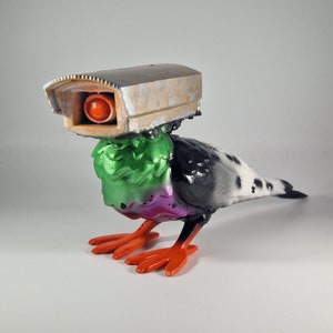 Puede incluir: Una escultura de pájaro blanco y negro con una cabeza verde y rosa y una cámara de vigilancia gris en la parte superior. El pájaro tiene patas naranjas.