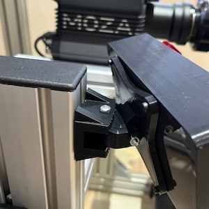 May include: Close-up of a black metal bracket attached to a silver aluminum rail. The bracket has a screw and a small black plastic piece. The bracket is attached to a black metal frame.