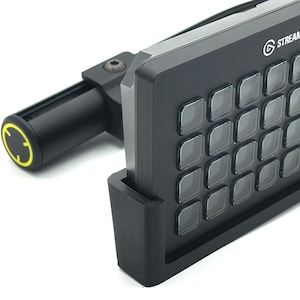 May include: A black Stream Deck with multiple square buttons and the Stream Deck logo. The device is mounted on a black stand and connected to a black arm with a yellow accent.