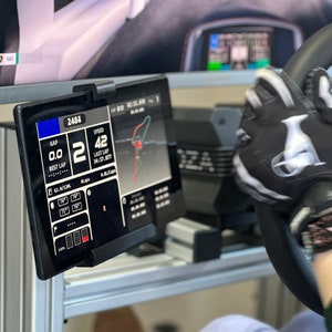 May include: A black racing simulator steering wheel with a black and white racing glove on it. The steering wheel has a black and white logo that says "MOZA". A black tablet with a white screen is mounted to the left of the steering wheel. The screen displays racing data, including speed, gap, and lap time. The text on the screen reads "2404", "SPEED 42", "GAP 0.0", "BEST LAP", "LAST LAP 34:37.877".