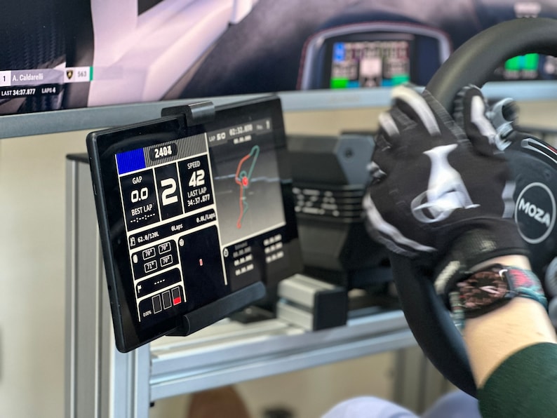 May include: A black racing simulator steering wheel with a black and white racing glove on it. The steering wheel has a black and white logo that says "MOZA". A black tablet with a white screen is mounted to the left of the steering wheel. The screen displays racing data, including speed, gap, and lap time. The text on the screen reads "2404", "SPEED 42", "GAP 0.0", "BEST LAP", "LAST LAP 34:37.877".