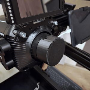 Può includere: Una fotocamera nera con un copriobiettivo. La fotocamera è montata su un treppiede nero con un piccolo monitor attaccato al lato.