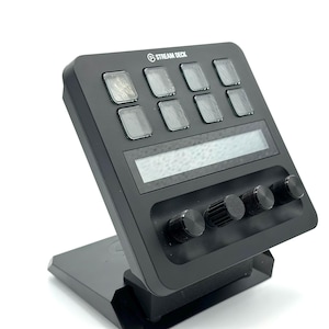 May include: A black Stream Deck device with eight illuminated buttons, a display screen, and four rotary knobs. The device is angled on a black stand, with the Stream Deck logo visible at the top.