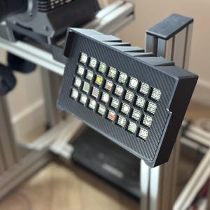 May include: A black, rectangular button box with a carbon fiber texture, featuring an array of illuminated buttons in various colors. The box is mounted on a silver metal frame, suggesting a sim racing or gaming setup.