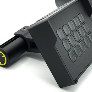 May include: A black device with a grid of clear buttons and a cylindrical handle. The handle has a yellow and black circular design. The device is angled, showing its various components and textures against a white background.