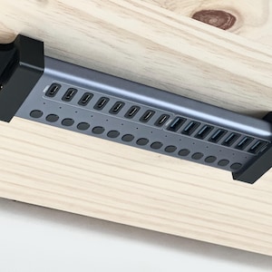 May include: A dark grey, rectangular USB charging hub mounted horizontally with black brackets. The device features a row of multiple USB ports and a corresponding row of circular buttons, secured with visible screws into a light wood surface.