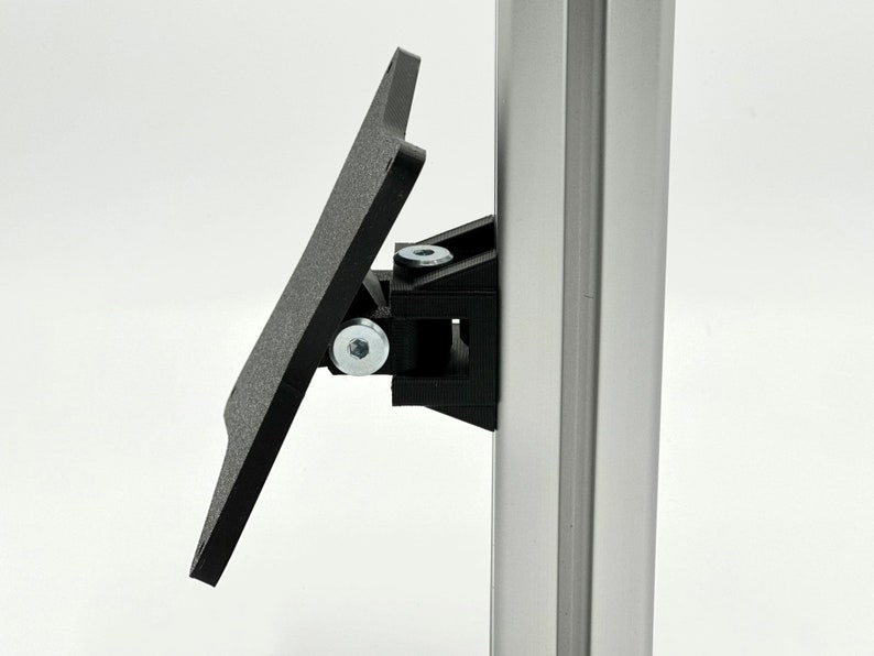 May include: A black plastic bracket with a flat, rectangular plate attached to it. The bracket is mounted to a silver metal pole.