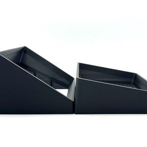 May include: Two black, angled storage trays are side-by-side against a white background. The trays have a rectangular base and a slanted top, creating a triangular shape. The trays are made of a matte black material.