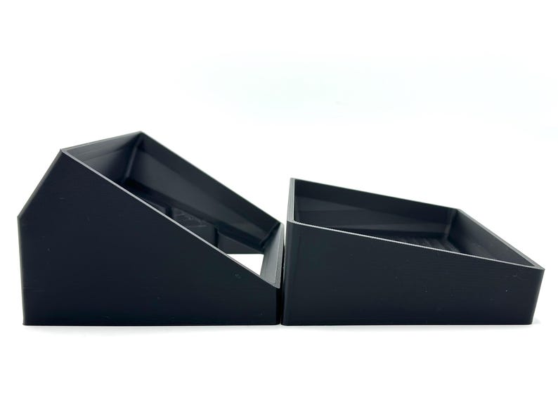 May include: Two black, angled storage trays are side-by-side against a white background. The trays have a rectangular base and a slanted top, creating a triangular shape. The trays are made of a matte black material.