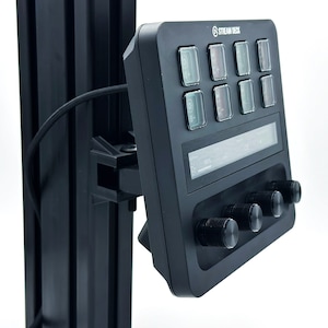 May include: A black Stream Deck with a screen and eight buttons. The buttons have red, green, and blue lights. There are three knobs on the bottom of the device. The Stream Deck is mounted on a black metal pole.
