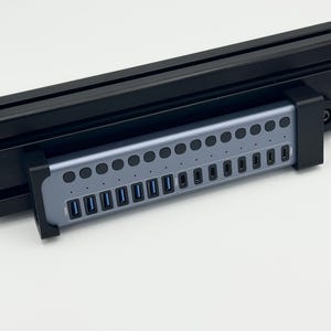 May include: A gray multi-port USB charging hub mounted in a black bracket. The device features a row of circular power buttons above a row of USB-C and USB-A ports. The text 100W is printed on the far left and far right ends of the hub face.