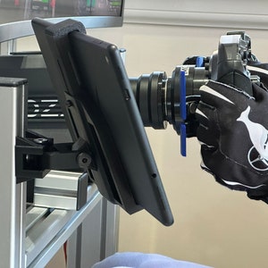 May include: A black tablet mounted on a silver stand with a black camera lens pointed towards the tablet. The camera is being held by a hand wearing a black glove with a white whale logo and the text "HIGH GROUND".