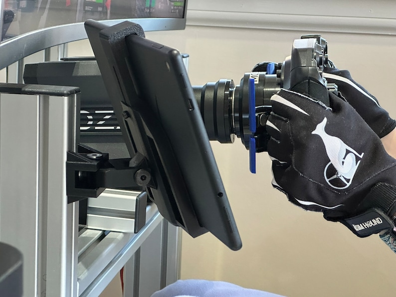 May include: A black tablet mounted on a silver stand with a black camera lens pointed towards the tablet. The camera is being held by a hand wearing a black glove with a white whale logo and the text "HIGH GROUND".