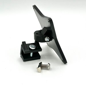 May include: A black plastic mount with a swivel joint and a clamp. The mount is designed to attach to a surface and hold a device, such as a camera or a phone.