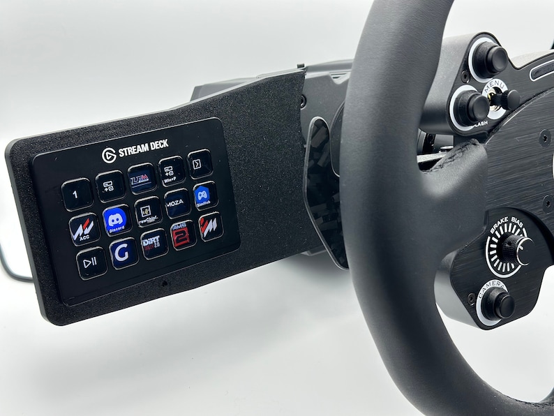 May include: A black racing wheel with a Stream Deck mounted on the left side. The Stream Deck has 15 buttons with various icons and text, including "G", "DIRT 2.0", and "MOZA". The wheel has a black leather grip, a brake knob, and a camera button.