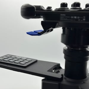 May include: A black camera mount with a control panel and a blue joystick. The mount has a keypad with 16 buttons.