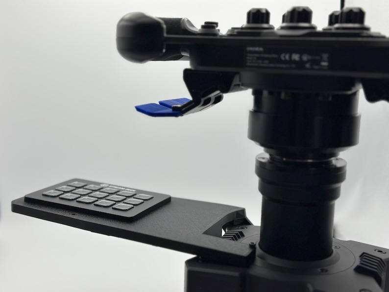 May include: A black camera mount with a control panel and a blue joystick. The mount has a keypad with 16 buttons.