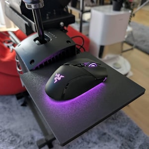 May include: A black gaming mouse with a purple underglow sits on a black surface. A black gear shift and a silver knob are also visible. The setup appears to be for a racing game.